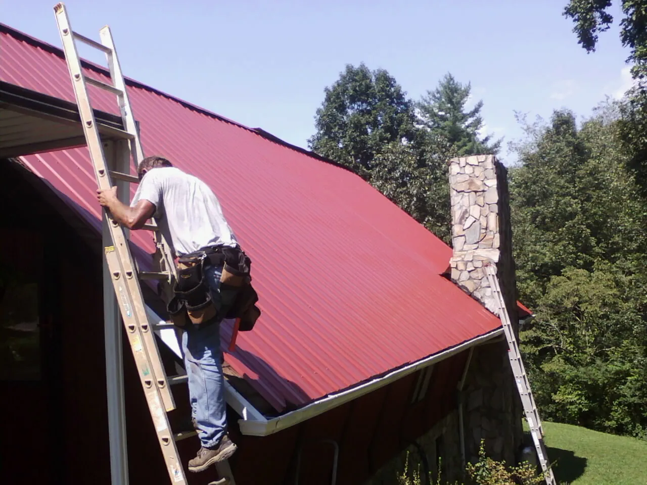 Roofer on a ladder beside a red metal roof and stone chimney — Soffit & Fascia Repair by Roofing Brothers in Homeworth